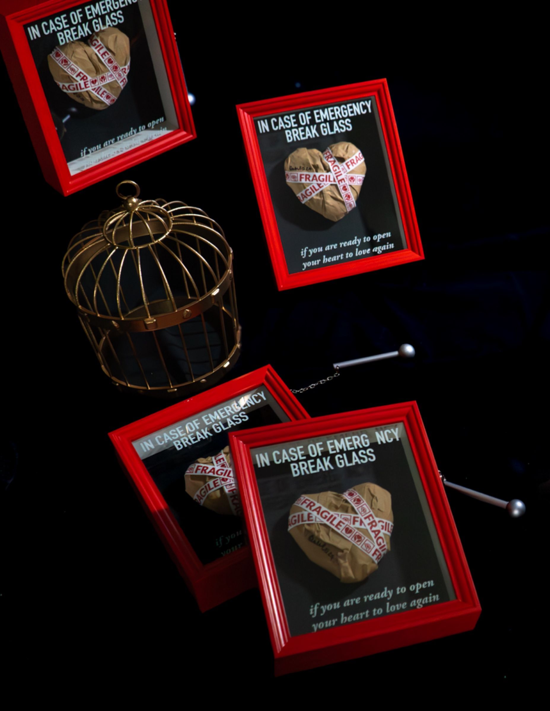Four red frames on a black ground, each holding a kraft paper heart wrapped in fragile tape under the words in case of emergency break glass, beside a golden cage.